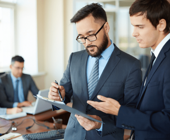 IT consulting services in Kochi – two business professionals in formal suits reviewing a project document on a clipboard in a modern office setting, discussing strategy and technology solutions.